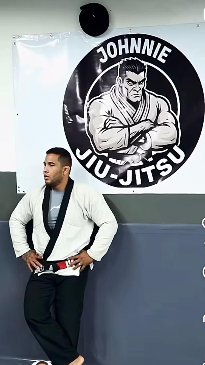 Professor Johnnie de kimono e faixa preta no tatame, em frente ao banner da Johnnie Jiu-Jitsu